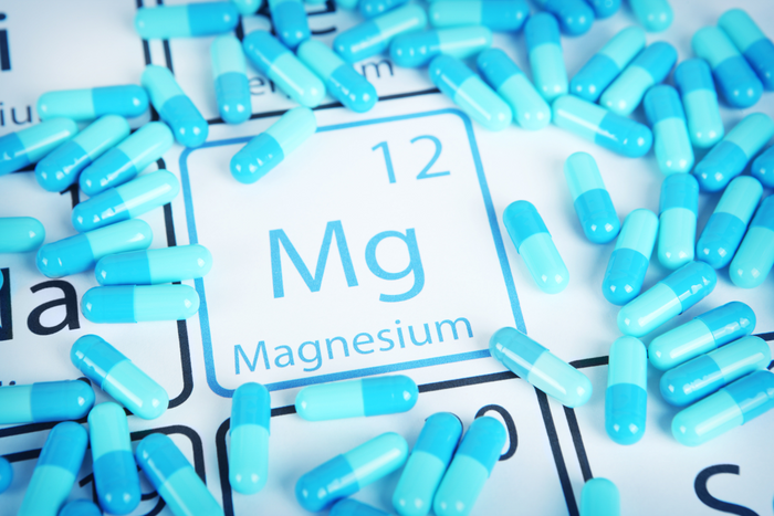 A close-up image showing a scatter of blue and white capsules around the periodic table symbol for Magnesium (Mg), which has an atomic number of 12, with the text