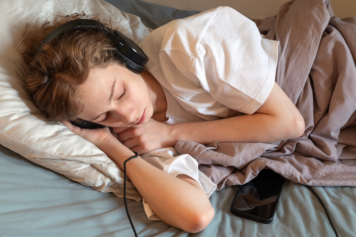 Music to fall asleep faster shown by person resting in bed with headphones and smartphone beside pillow