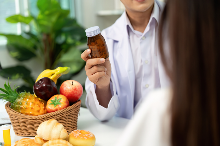 A person in a white coat holds up a small brown bottle while discussing a basket of fruit and pastries with someone across a table.
