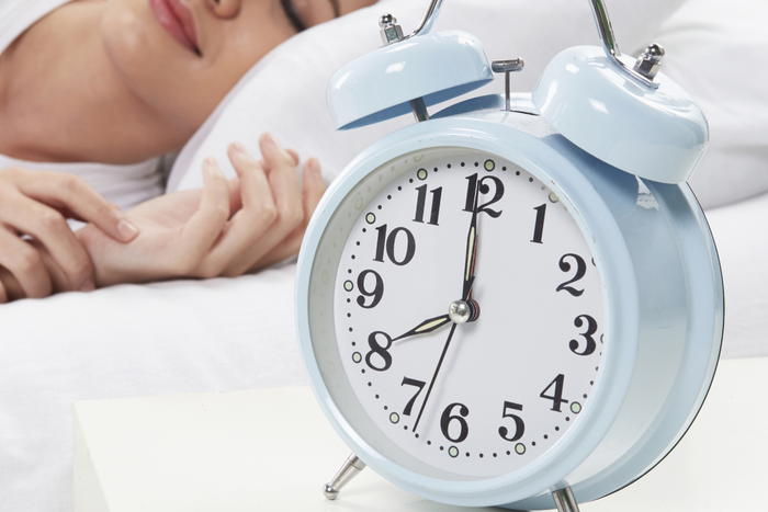 A light blue vintage alarm clock on a white nightstand, with a woman sleeping peacefully in the blurred background.