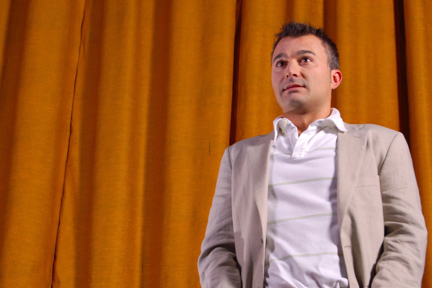A man in a light brown jacket and white collared shirt stands in front of an orange curtain.