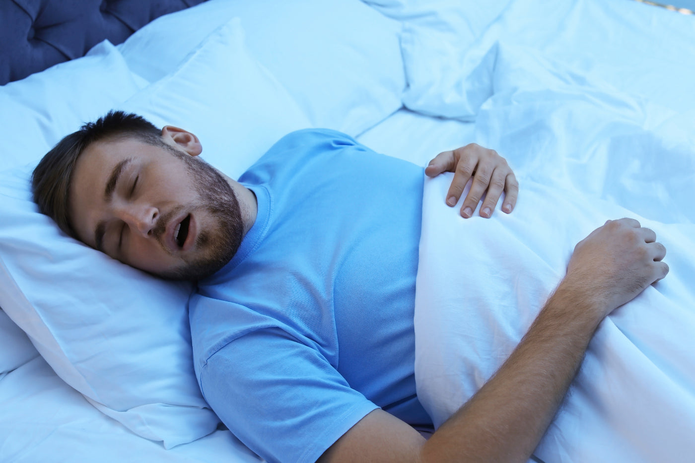 A bearded man in a blue t-shirt sleeps deeply on his back with his mouth slightly open.