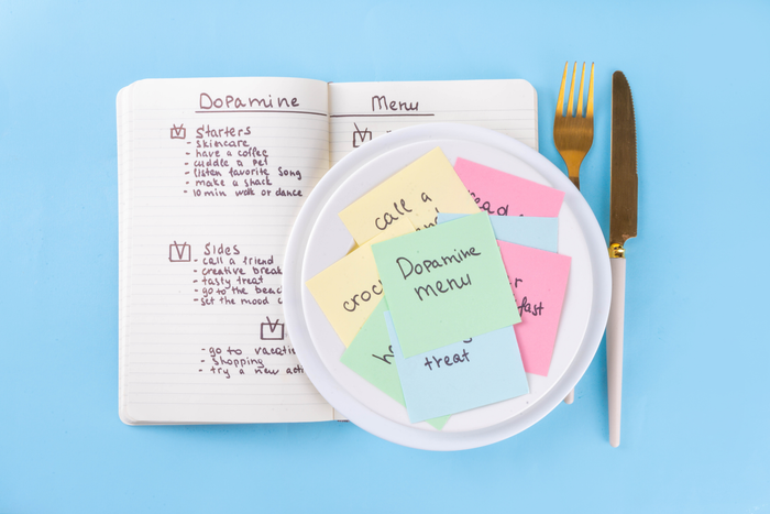 A notebook open to a Dopamine menu with starters and sides listed. A plate with colorful sticky notes and gold cutlery sits on a blue background.