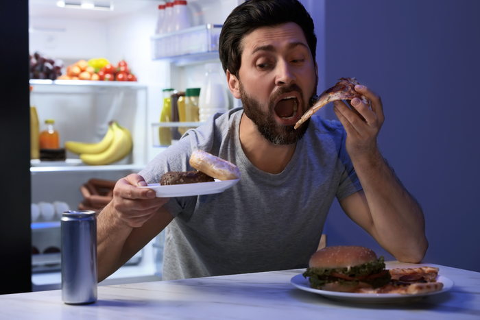 A person is eating pizza in front of an open refrigerator with a plate of food and a can of soda nearby.