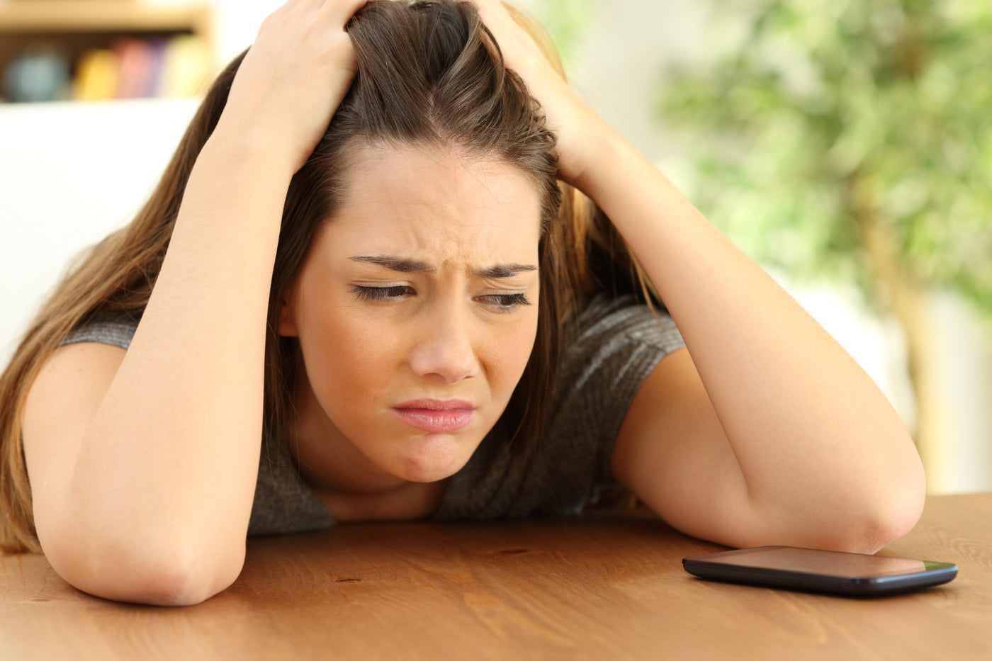 A person with hands in their hair leans over a wooden table with a cell phone.