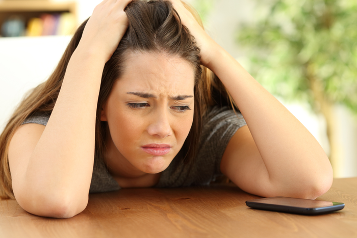 A person with hands in their hair leans over a wooden table with a cell phone.