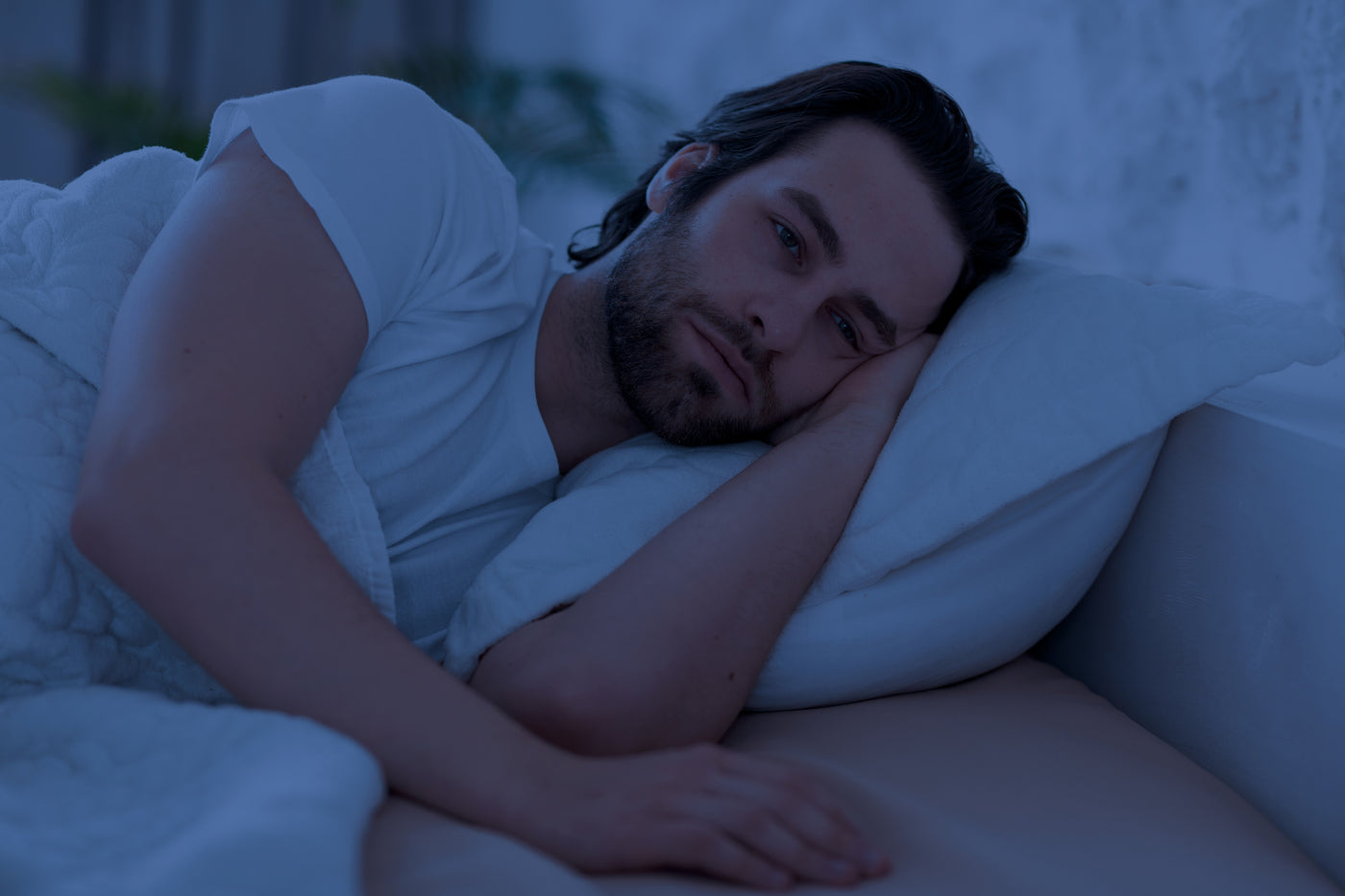 Man lying awake in bed under cool blue nighttime light, illustrating sleeplessness and delayed bedtime