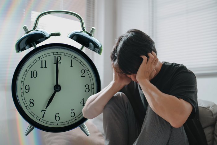 A stressed man sitting in bed holding his head in distress, superimposed with a large alarm clock showing seven o'clock.
