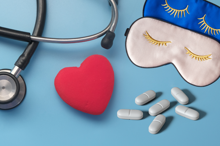 A stethoscope placed next to a bright red heart shape and a small cluster of white pills and sleeping masks on a light blue background.