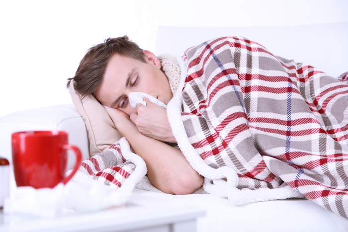 A tired man wrapped in a plaid blanket resting on a sofa while holding a tissue to his nose, with a red mug of hot tea in the foreground.