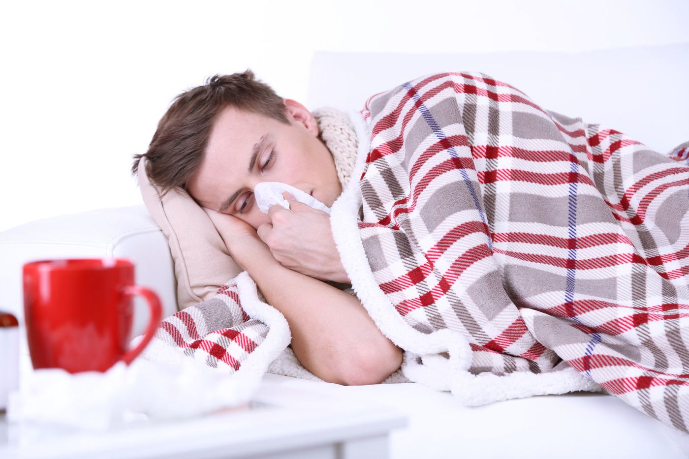 A tired man wrapped in a plaid blanket resting on a sofa while holding a tissue to his nose, with a red mug of hot tea in the foreground.