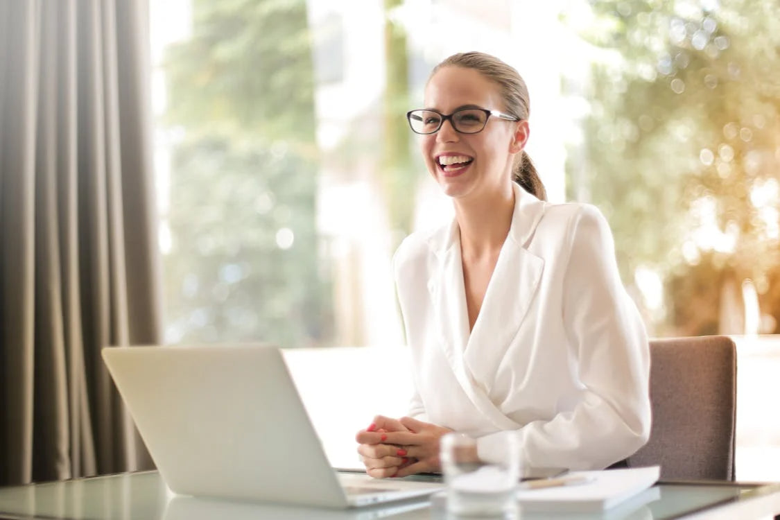 A smiling woman with glasses and a white blazer sits at a desk with a laptop and a glass of water.