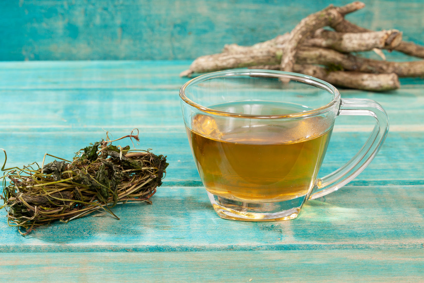 A clear glass mug of amber-colored tea sits on a turquoise wooden table next to a bundle of dried valerian root.