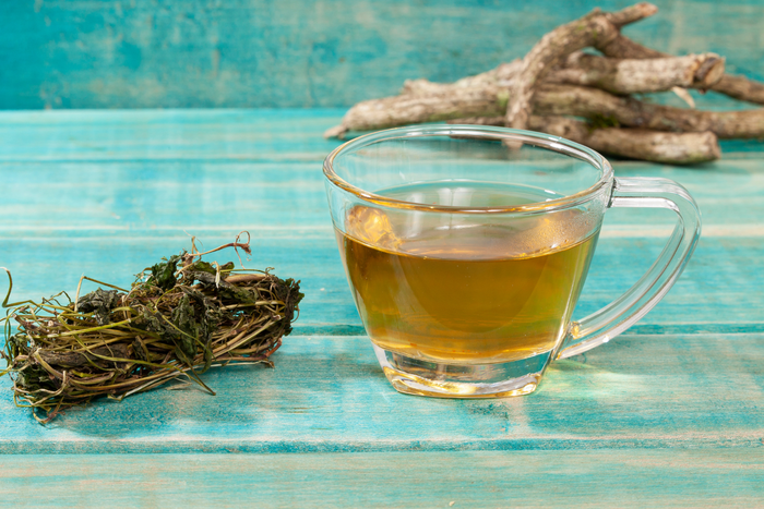 A clear glass mug of amber-colored tea sits on a turquoise wooden table next to a bundle of dried valerian root.