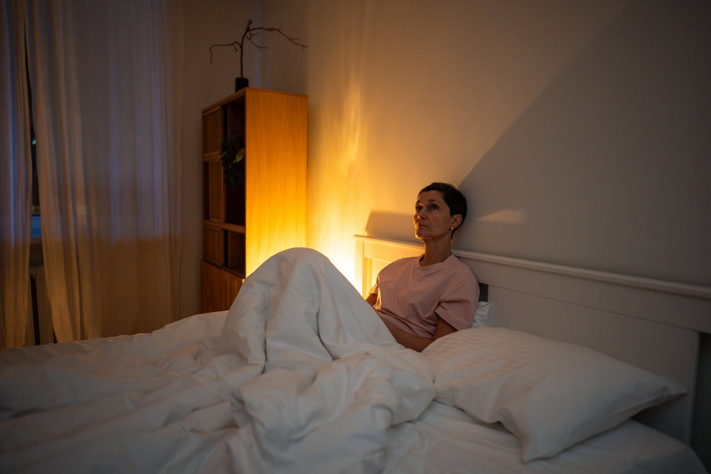 Woman sitting awake in bed at night with warm light, showing stress and overthinking before sleep